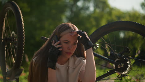 joven sentada al lado de su bicicleta al revés, haciendo una llamada telefónica con una expresión cansada e infeliz, reflejando su estado de ánimo mientras está rodeada de exuberante vegetación y árboles