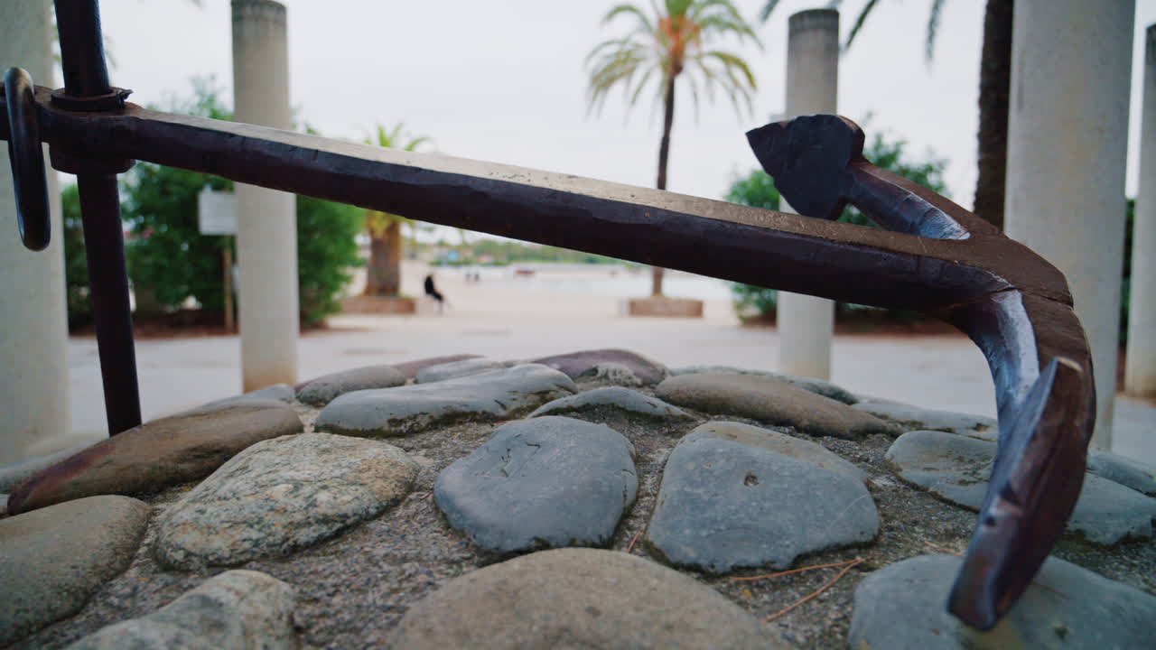 A metal anchor displayed on a stone base near palm trees and a seaside promenade