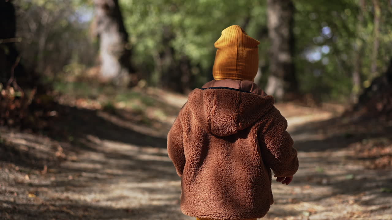 Following the lovely baby boy wearing brown jacket and yellow cap. Sweet kid on the walk in the autumn park.