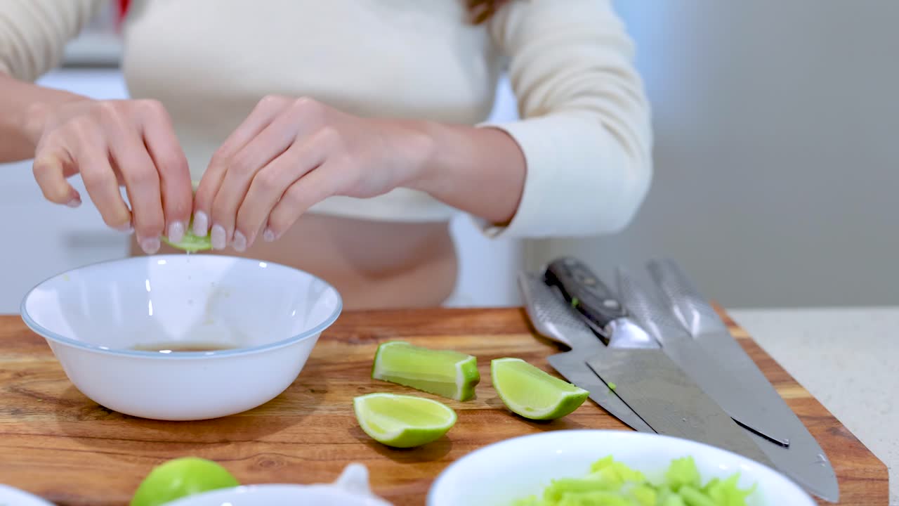 Hands squeeze lime halves into a bowl on a wooden cutting board, surrounded by kitchen utensils and lime pieces