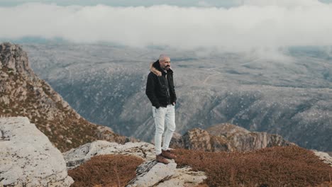 Ein-Gutaussehender-Mann-Mit-Kurzen-Haaren-Und-Schnurrbart-Steht-Auf-Einem-Felsen,-Die-Hände-In-Den-Taschen,-Und-überblickt-Die-Atemberaubende-Berglandschaft-Mit-Nebligen-Wolken-Am-Horizont