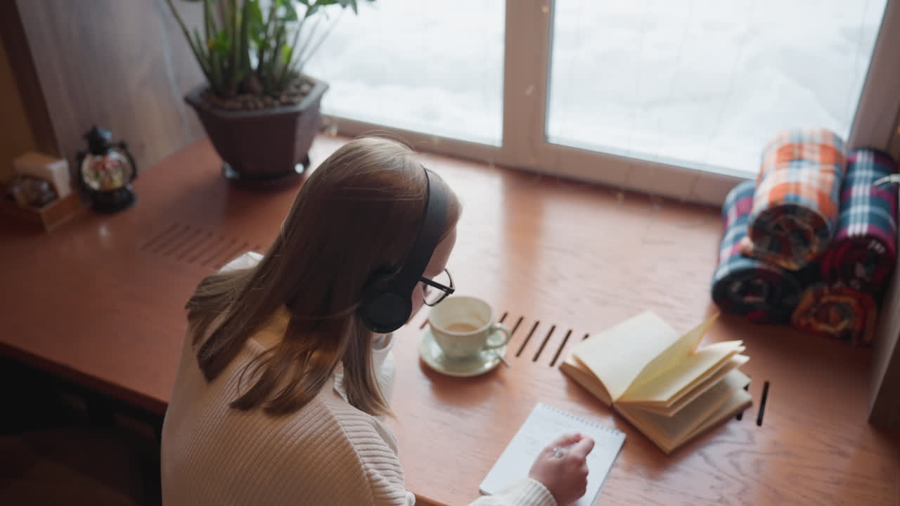 overhead view of student in white sweater wearing headphone writing in notebook while studying open book near window with warm drink and folded cloth on wooden table