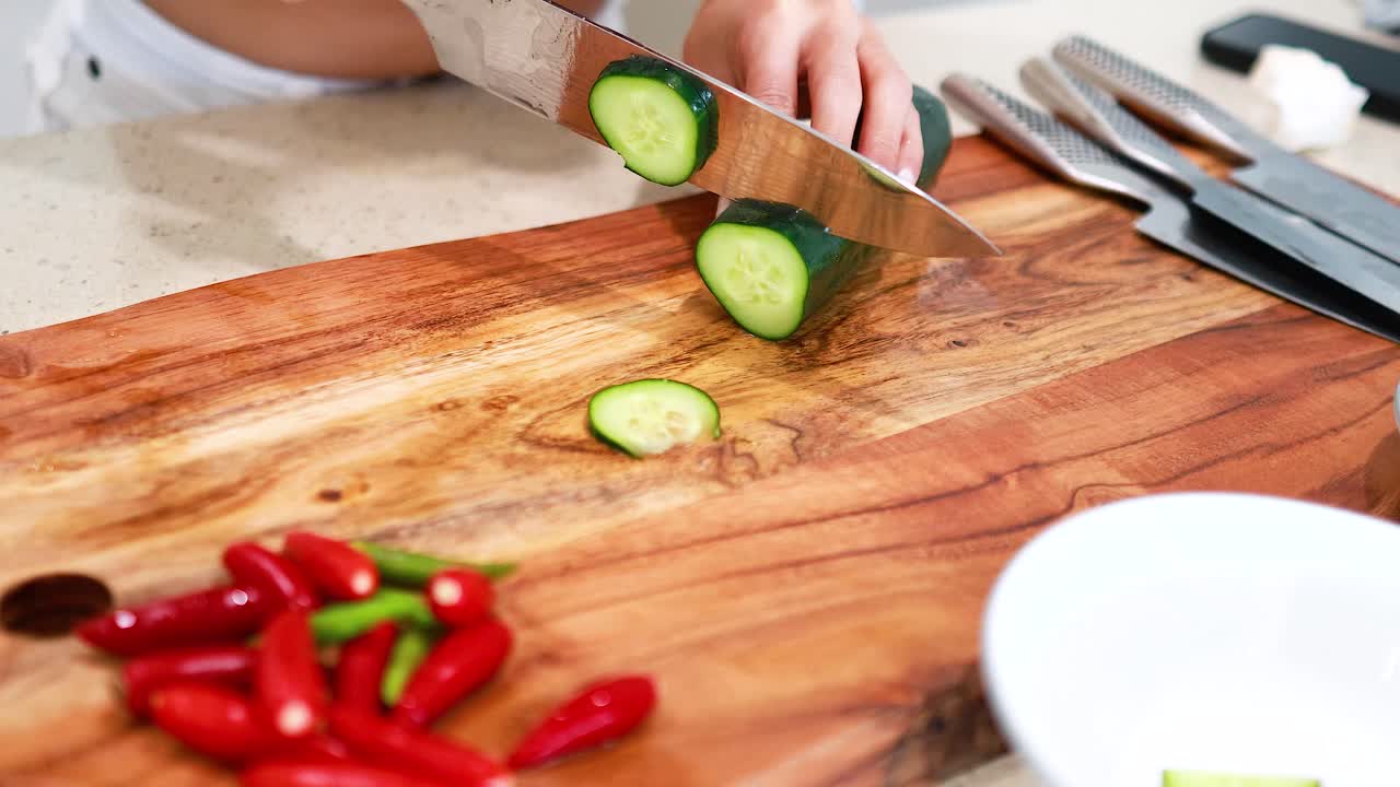 Hands slicing cucumber on a wooden board with vibrant lighting, showcasing culinary preparation in a modern kitchen setting
