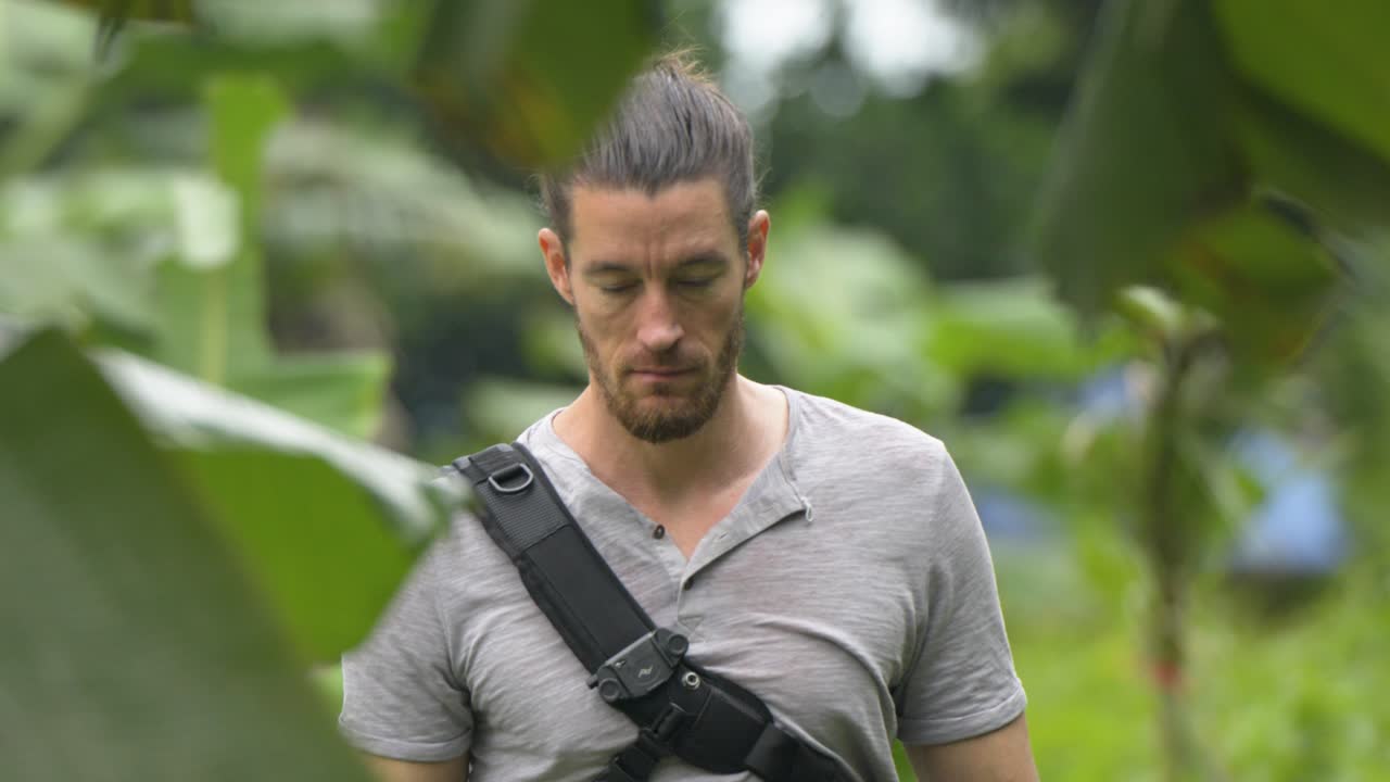 close-up shot of a Caucasian man with tied-back hair walking straight ahead, facing the camera and looking around, in a tropical forest with banana trees