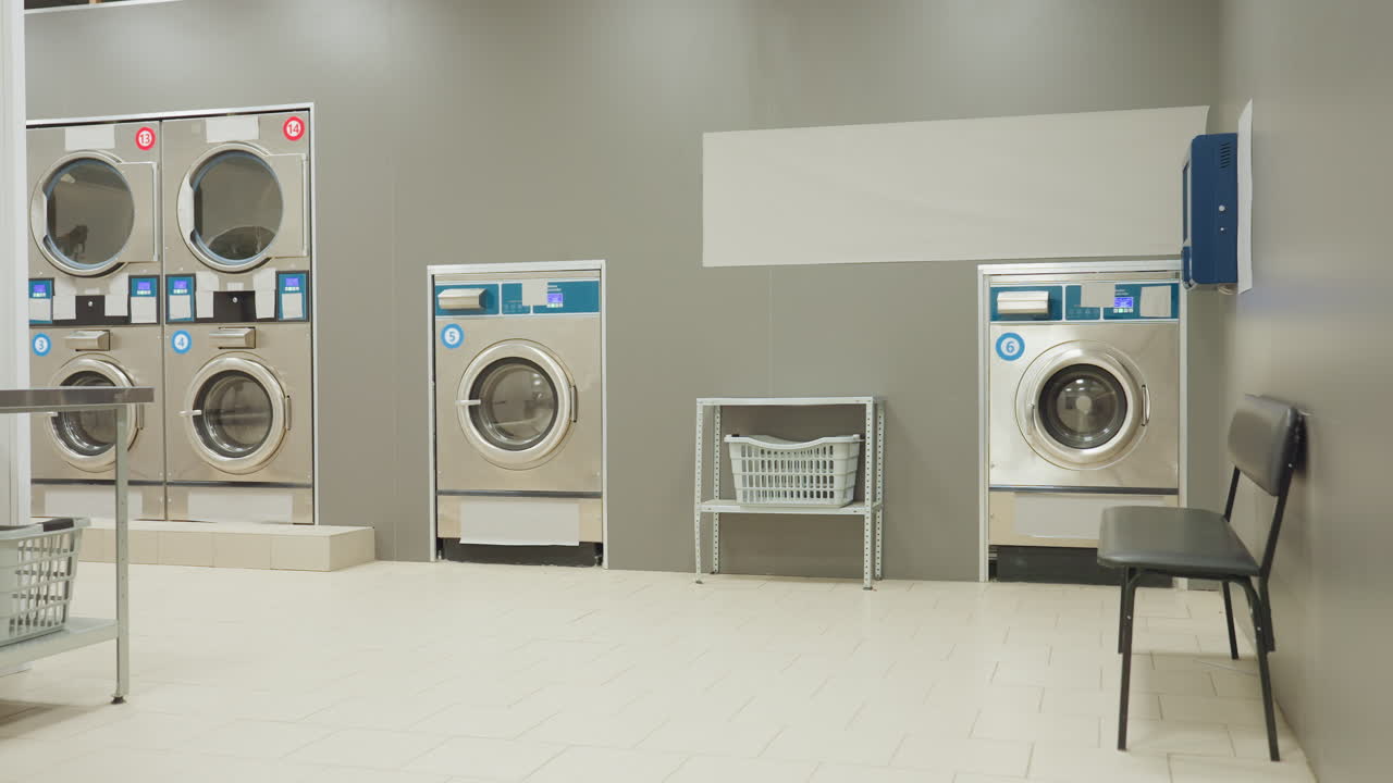 Dim laundry shop interior with metallic washing machines lined against gray wall, laundry basket shelf, and black bench in low light setting as digital displays glow faintly creating moody atmosphere