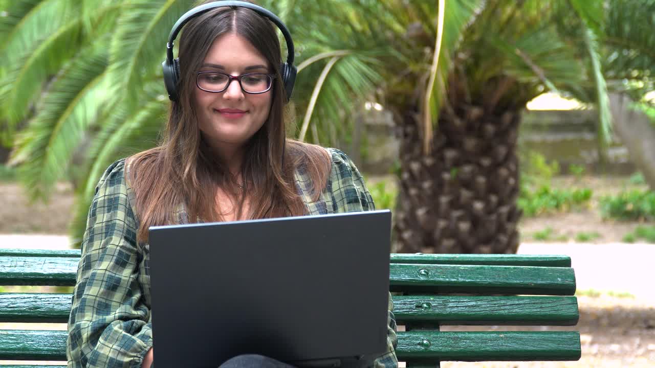 bella estudiante con auriculares trabajando en una laptop en un banco en el parque