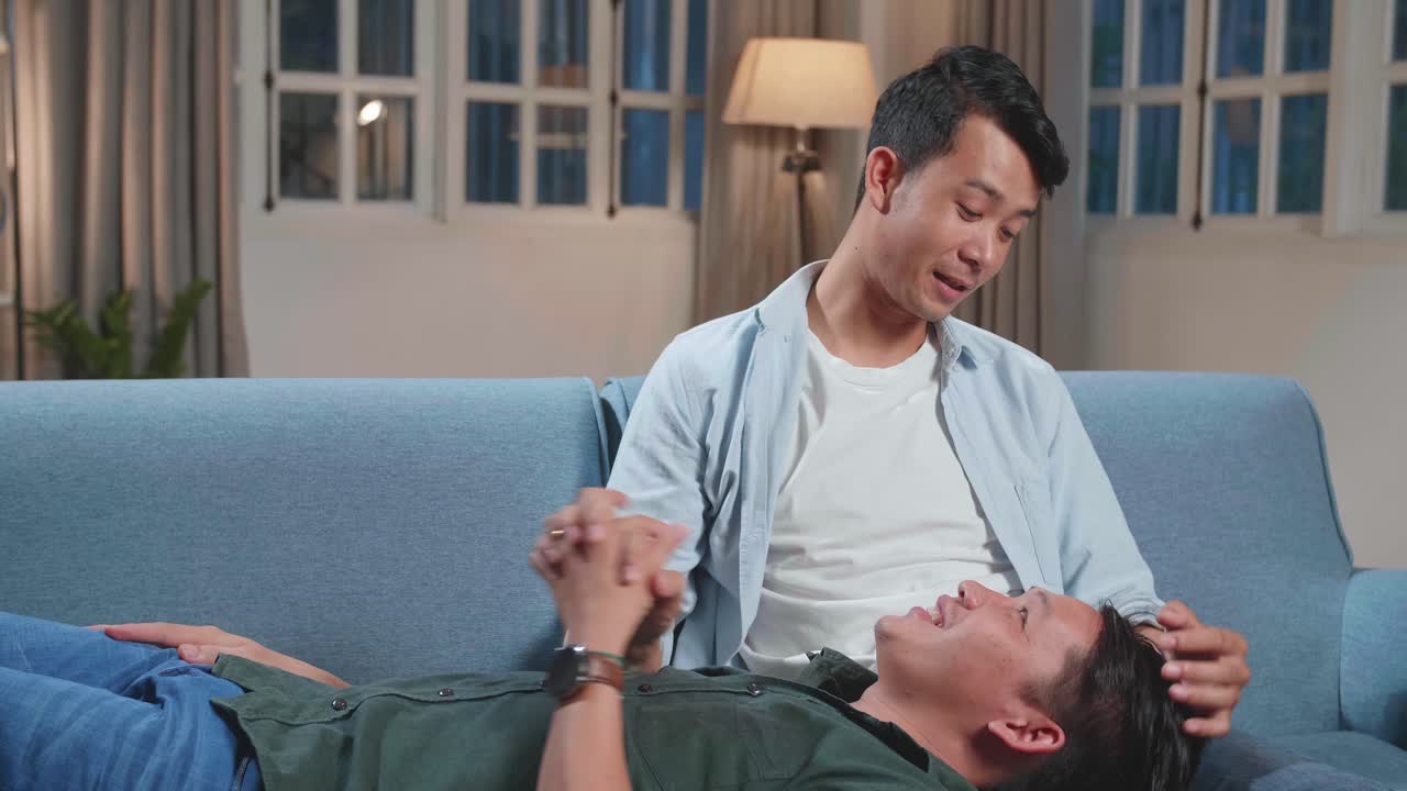 Young Asian Gay Couple Spend Time At Home. Two Happy Man In Love Lie Down On Sofa And Cuddle In Casual Clothes. Cute Lgbt Relationship Content