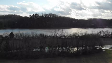 Aerial-footage-coming-down-over-the-park-while-man-is-biking-and-dog-is-running-river-in-background