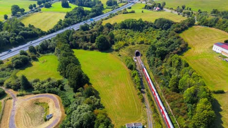 passenger train exiting tunnel through green countryside near highway from aerial drone view