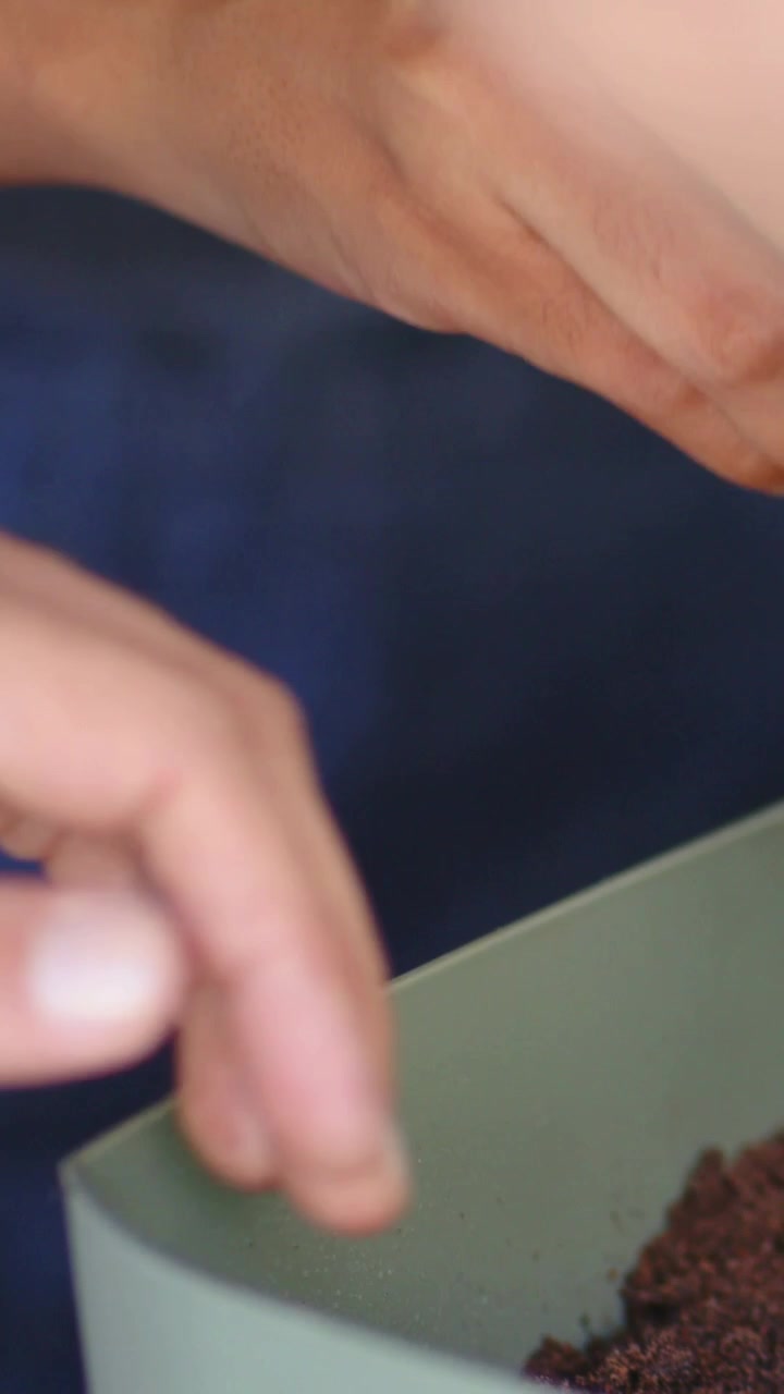 Vertical video: Tilting pink scoop, hands adding soil into matte green-gray planter, blue fabric