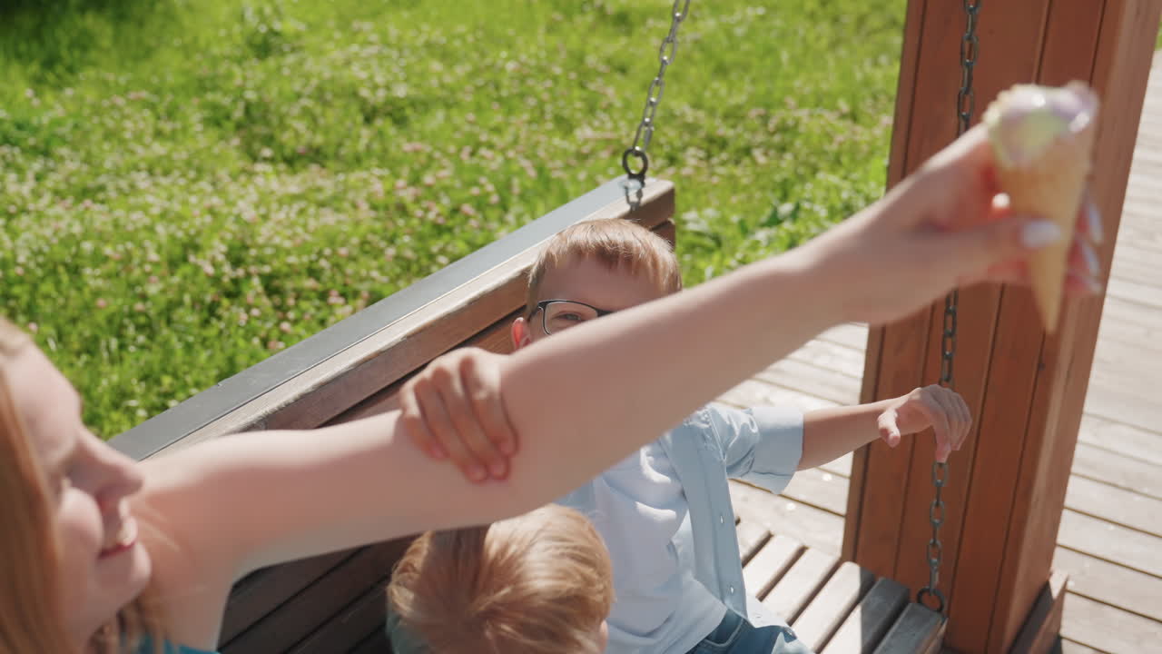 Wit gezin parkweekend, oudere jongen reikt speels om een ijsje uit moeders hand te pakken, zacht trekken aan de arm terwijl ze op een schommelbank zitten, zonlicht op het gras, levendige broer-zus interactie en oprechte vreugde.