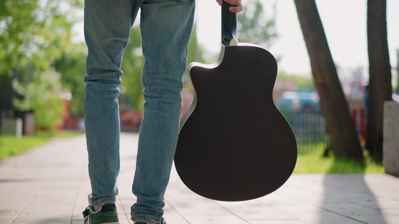 Guitarrista caminando por un sendero soleado en un parque, con una funda de guitarra acústica, en vaqueros y zapatillas. Primer plano de la parte inferior del cuerpo y la sombra en el pavimento, árboles frondosos y valla verde al fondo, paso informal.