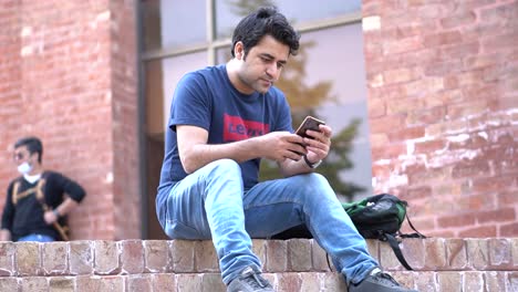 a university student man sitting on the stairs on a sunny day using his smartphone while people pass by.  men using smartphones outdoor