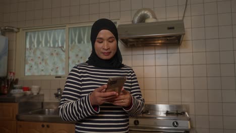 Mujer-Musulmana-Asiática-Indonesia-Con-Hijab-Enviando-Mensajes-De-Texto-Con-Teléfono-En-La-Cocina