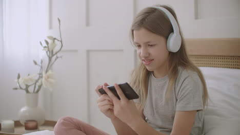 little girl is using smartphone and wireless headphones sitting on bed in room playing game and smiling