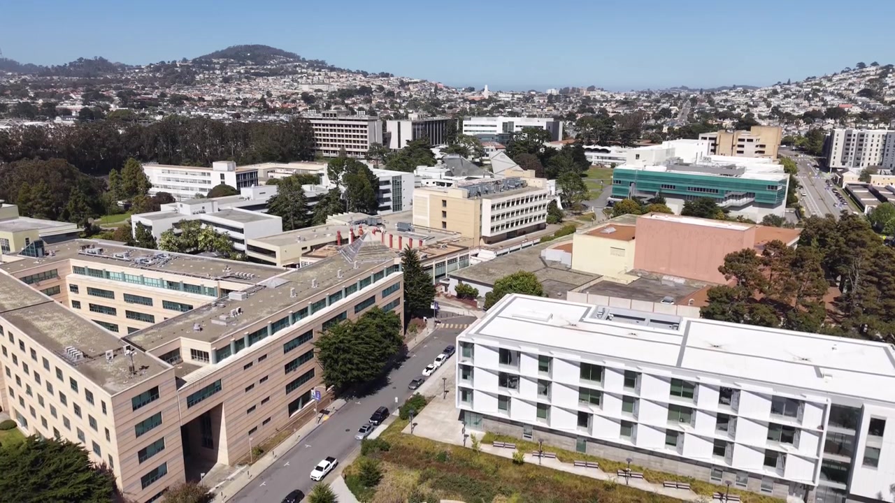 Imagens aéreas do campus da Universidade Estadual de São Francisco, apresentando arquitetura moderna e espaços verdes exuberantes sob um céu azul claro, com a cidade circundante e colinas ao fundo