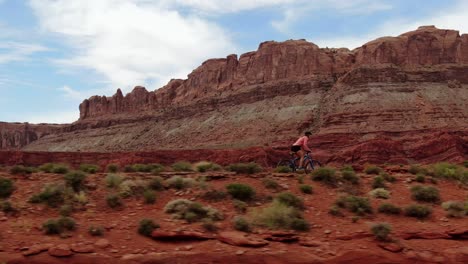 aerial shot following woman riding mountain bike in red moab desert with mountains