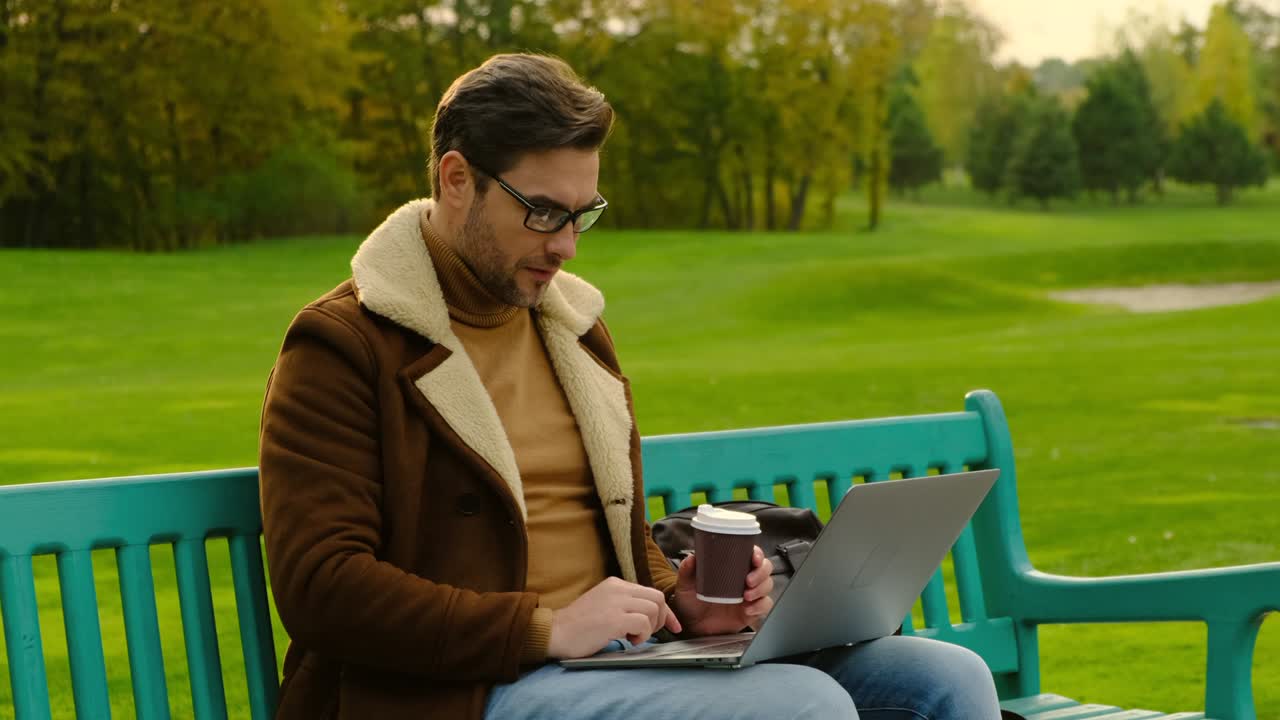 stylish man in a brown jacket sits on a park bench with a laptop on the Park