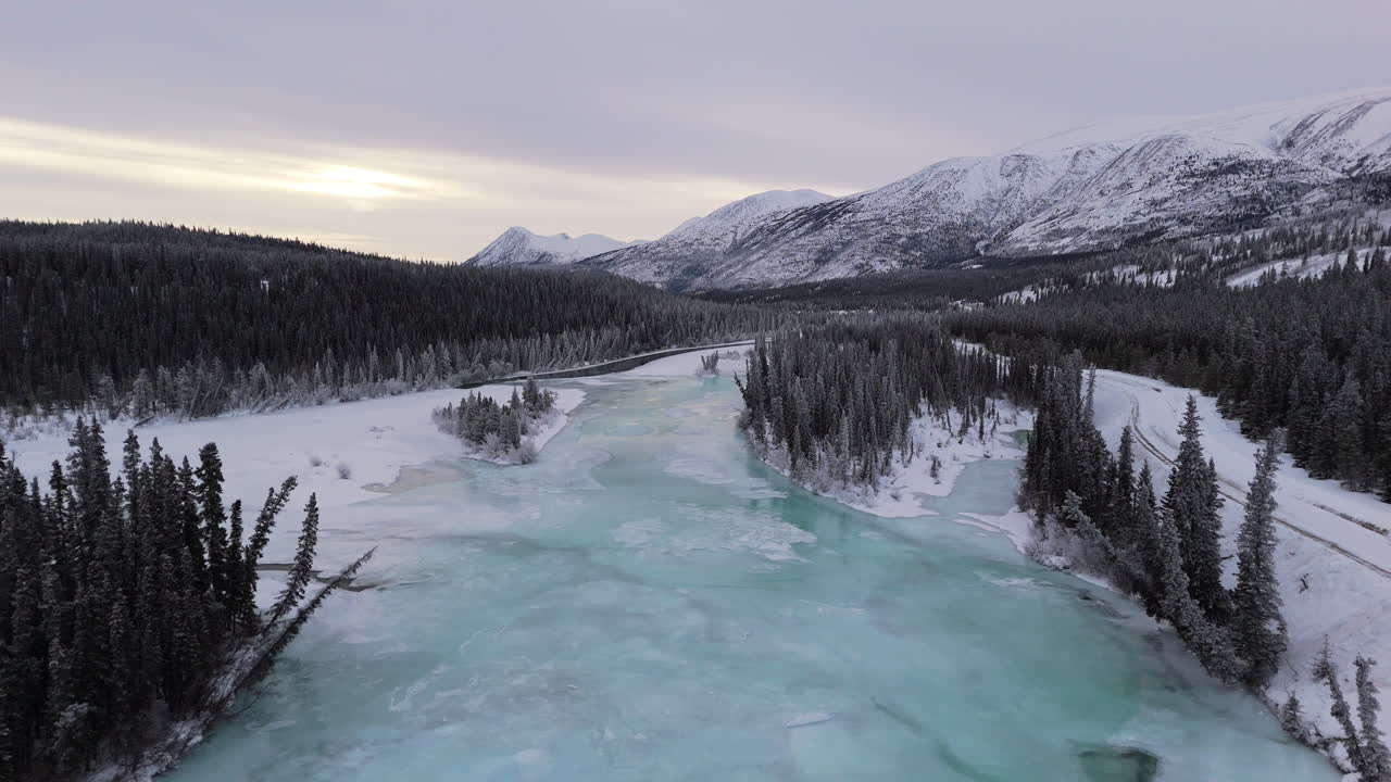 A smooth aerial dolly forward shot travels over icy trees and the frozen Takhini River with a majestic mountain backdrop. This dynamic footage captures the pristine winter landscape of Yukon
