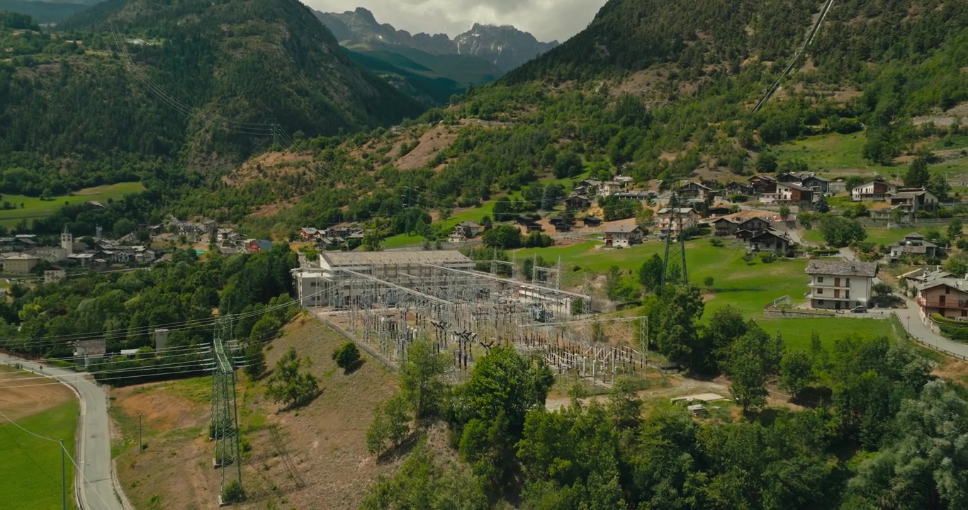Dron se blíží k rozvodně vysokého napětí s mnoha elektrickými vedeními v horské vesnici, zdůrazňující moderní energetickou infrastrukturu v alpské krajině.