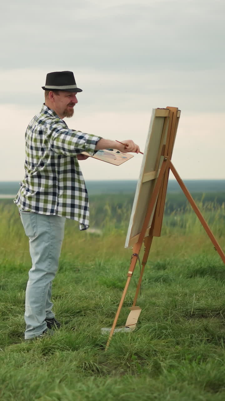 검은 모자, 체크색 셔츠, 청바지를 입은 예술가는 조용한 잔디에 설치된 큰 캔버스를 그리는 데 깊이 집중하고 있습니다. 조용한 풍경은 창의적인 과정을 향상시니다.