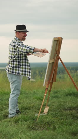 un artista con un sombrero negro, camisa a cuadros y vaqueros está profundamente enfocado en pintar un gran lienzo establecido en un campo de hierba sereno. el paisaje tranquilo mejora el proceso creativo