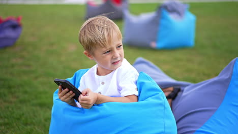 cute fair-haired little boy lies in bean bag chair. blue-eyed kid looks at phone sometimes distracting. close up. blurred backdrop.