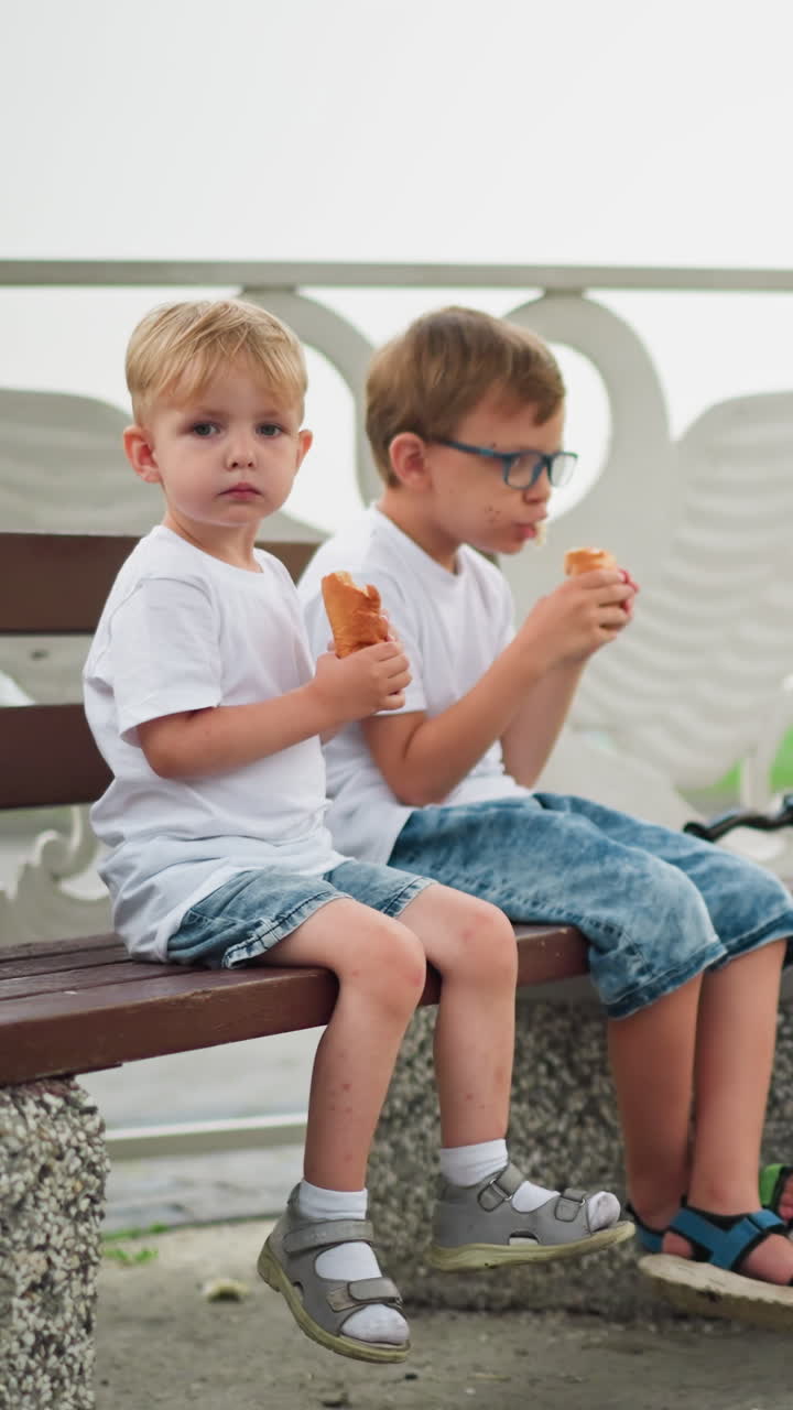 двое молодых братьев и сестер сидят вместе на скамейке, старший ребенок ест колбасу, в то время как младший держит в центре внимания, скутер находится рядом