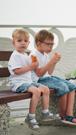 dos hermanos jóvenes se sientan juntos en un banco, el niño mayor está comiendo un rollo de salchichas, mientras que el más joven está manteniendo la mirada enfocada, un scooter está cerca