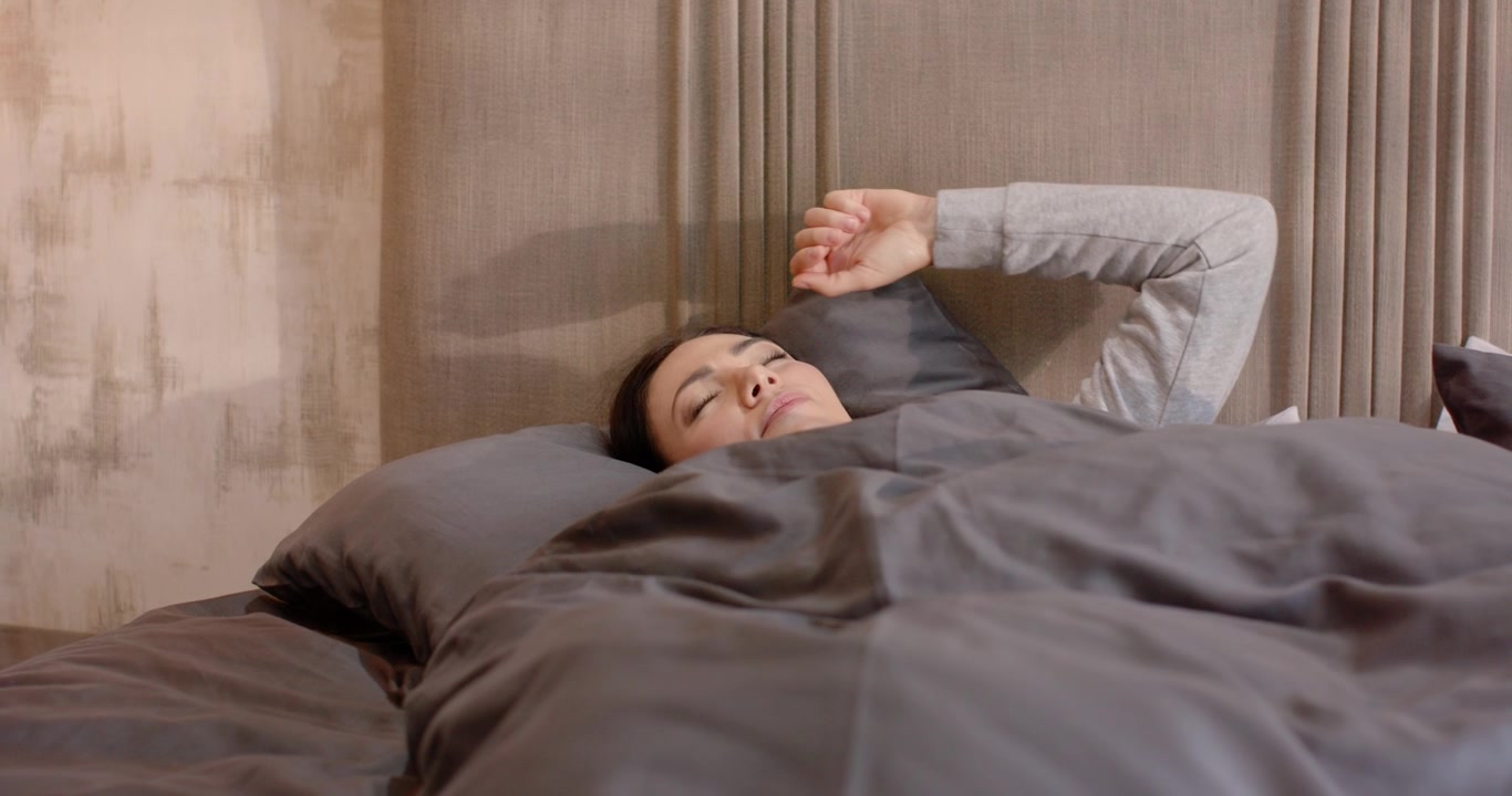 Wanita dewasa bangun tidur, berbaring di tempat tidur di bawah selimut abu-abu di samping sandaran kepala berlapis panel, membuka mata, menggosok mata