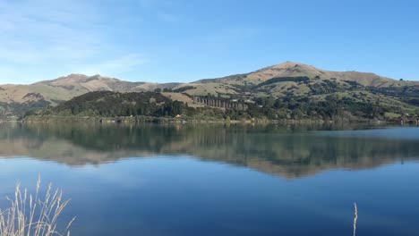 Ripples-of-water-flow-slowly-across-reflection-of-hills-on-a-clear,-calm-morning---French-Farm-Bay,-Akaroa-Harbor