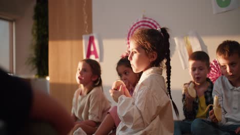 A-little-girl-with-a-braided-hairstyle-in-a-white-shirt-eats-an-apple-at-recess-and-during-lunch-in-a-club-for-preparing-preschool-children-for-school