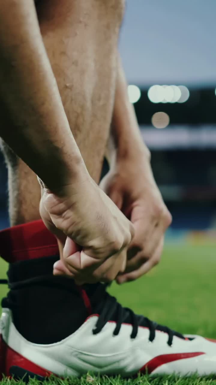 Video verticale: giocatore accovacciato che si allaccia i lacci bianchi o rossi delle scarpe da calcio sull'erba, in tenuta rossa e pronto per la partita
