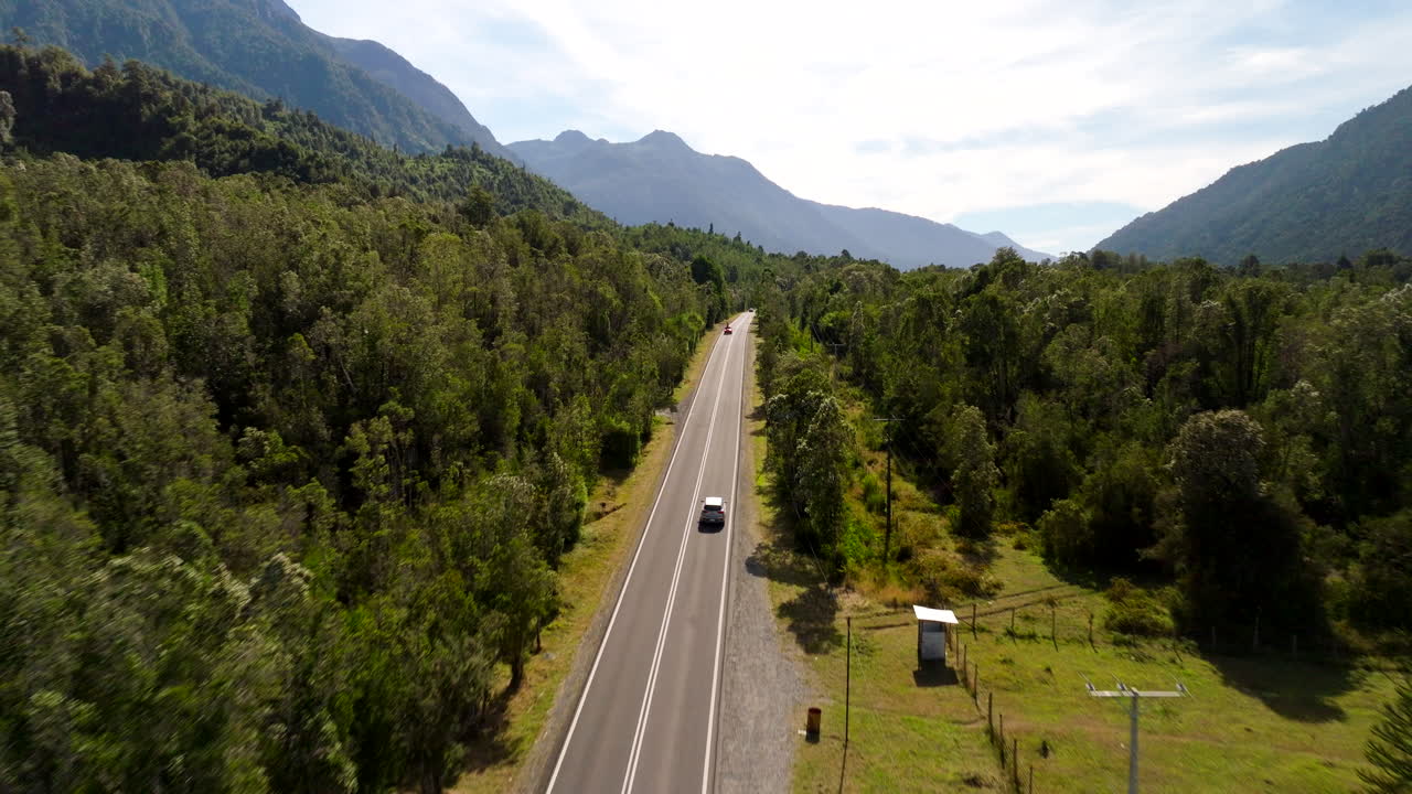 Aerial drone view of a car driving along a paved road surrounded by dense green forest and mountains near the Petrohue River in southern Chile. Ideal for travel and road trip concepts