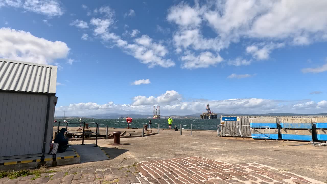 Several individuals in high-visibility jackets walk along a stone pier by the sea under bright daylight, with boats and a lighthouse visible in the distance. The camera pans slowly, capturing the coastal environment and dynamic sky