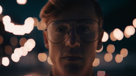 retrato joven pelirrojo con gafas en la azotea por la noche mirando una expresión seria con hermosas luces de fiesta en el fondo del concepto de juventud urbana