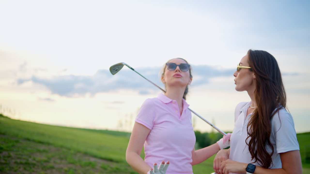 Two women dressed in white and pink clothes, holding golf clubs and talking on the course