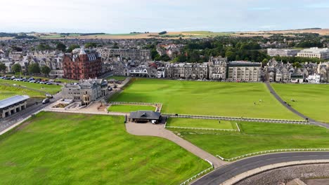 drone camera glides above green fairways and historic brick buildings at st andrews, scotland, under bright daylight with smooth, forward motion