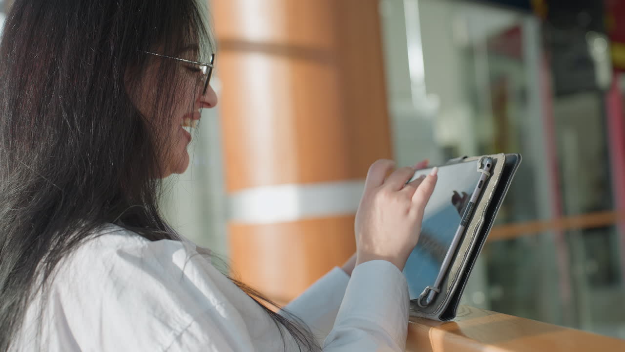 Side view of woman with long dark hair and glasses smiling while using digital tablet against sleek interior background with glass railing and warm wooden columns under natural light