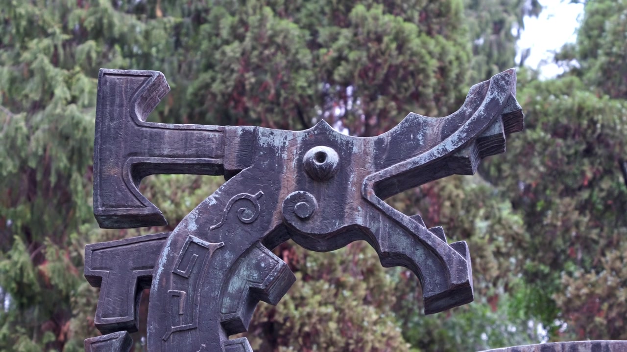Yemyeşil ağaçların fonunda karmaşık geometrik desenlere sahip bronz bir heykel. Kültürel belgeseller veya sanat çalışmaları için idealdir