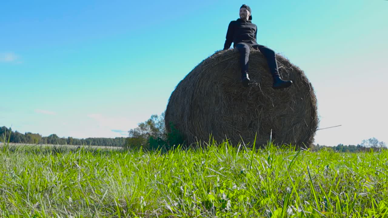 Person with black clothing sitting on top of round or rounded hay bale and moving his feet in slow motion that makes some hay fall. Man is happy and looks and watches around while sun is shining.