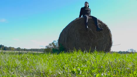 person with black clothing sitting on top of round or rounded hay bale and moving his feet in slow motion that makes some hay fall. man is happy and looks and watches around while sun is shining.