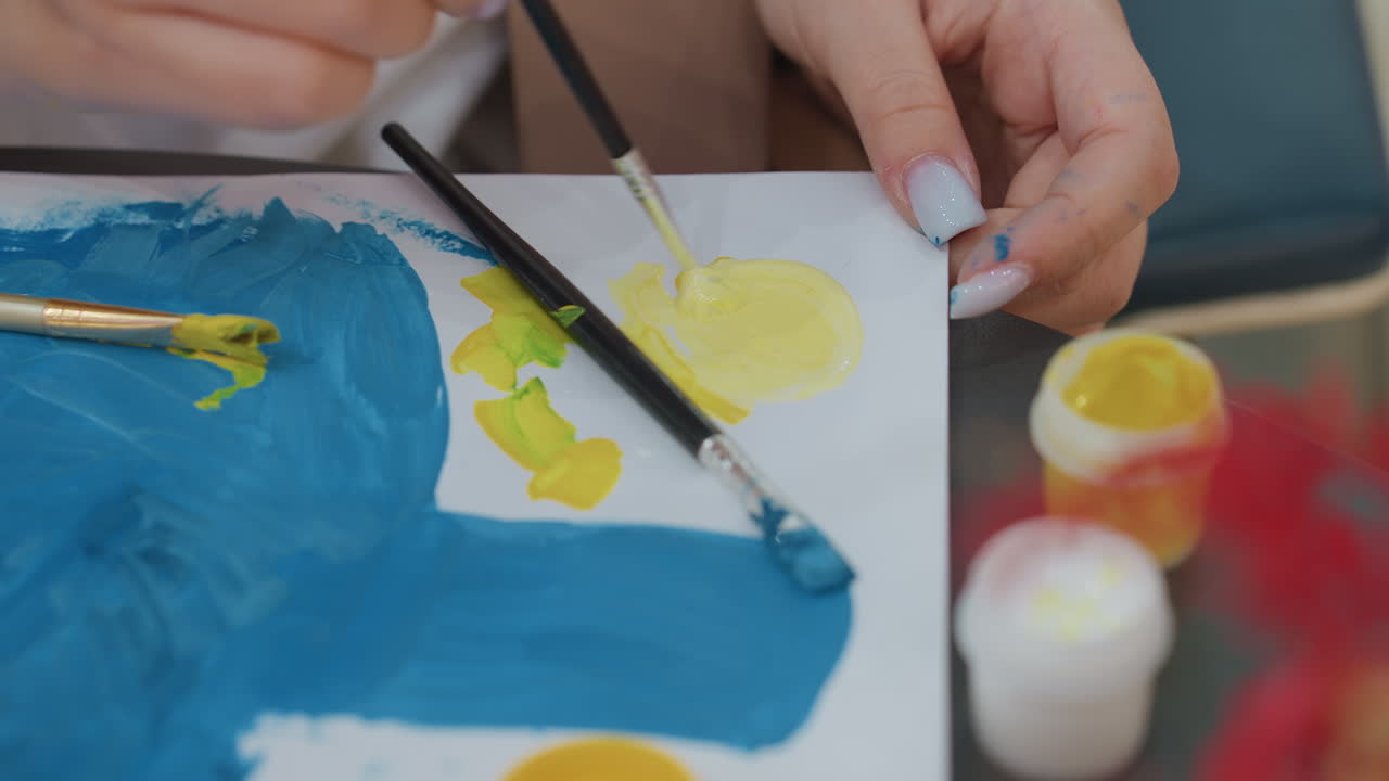클로즈업된 흰색 여성의 손이 캔버스 위에 노란색 페인트를 섞고 있으며, 아래에는 파란색 밑칠이 되어 있어요. 테이블 위에는 붓과 병이 있고, 손길 하나하나에 집중하며 세심하게 칠하고 있어요. 깔끔한 매니큐어가 눈에 띄고, 친밀하고 창의적인 분위기가 느껴져요.