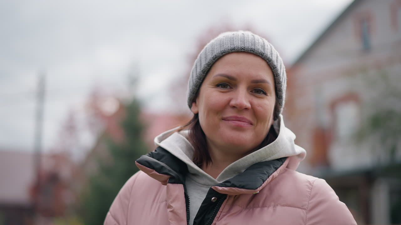 분홍색 재과 회색 모자를 입은 행복한 여성이 카메라에 따뜻하게 미소 짓고, 활발한 가을 잎자루, 집 및 정원을 특징으로하는 흐릿한 배경에 대 한 야외에 서 있습니다.