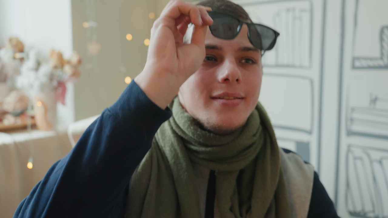 College student flips sunglasses with grin beside illustrated bookshelf, olive scarf and soft window lighting, candid movement and charming gesture, relaxed vibe for lifestyle and vlog content showing