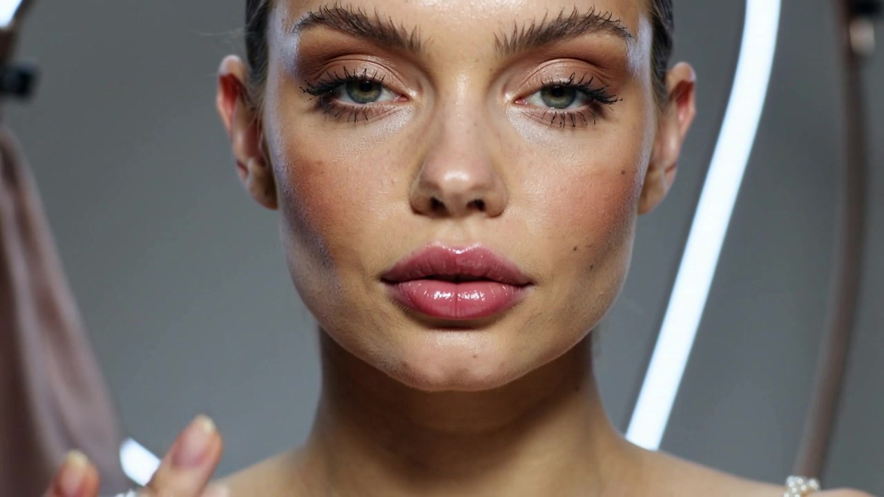 Touching rings to lower lip, thin-strapped model blending gloss in studio, LED arc, showing look