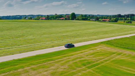 Sideway-drone-shot-of-a-blue-BMW-car-driving-down-a-scenic-road-surrounded-by-lush-green-meadows-on-a-sunny-warm-summer-day