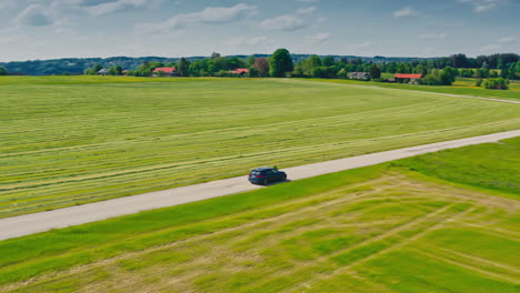 Seitliche-Drohnenaufnahme-Eines-Blauen-BMW-Autos,-Das-An-Einem-Sonnigen,-Warmen-Sommertag-Eine-Malerische-Straße-Entlang-Fährt,-Umgeben-Von-üppigen-Grünen-Wiesen