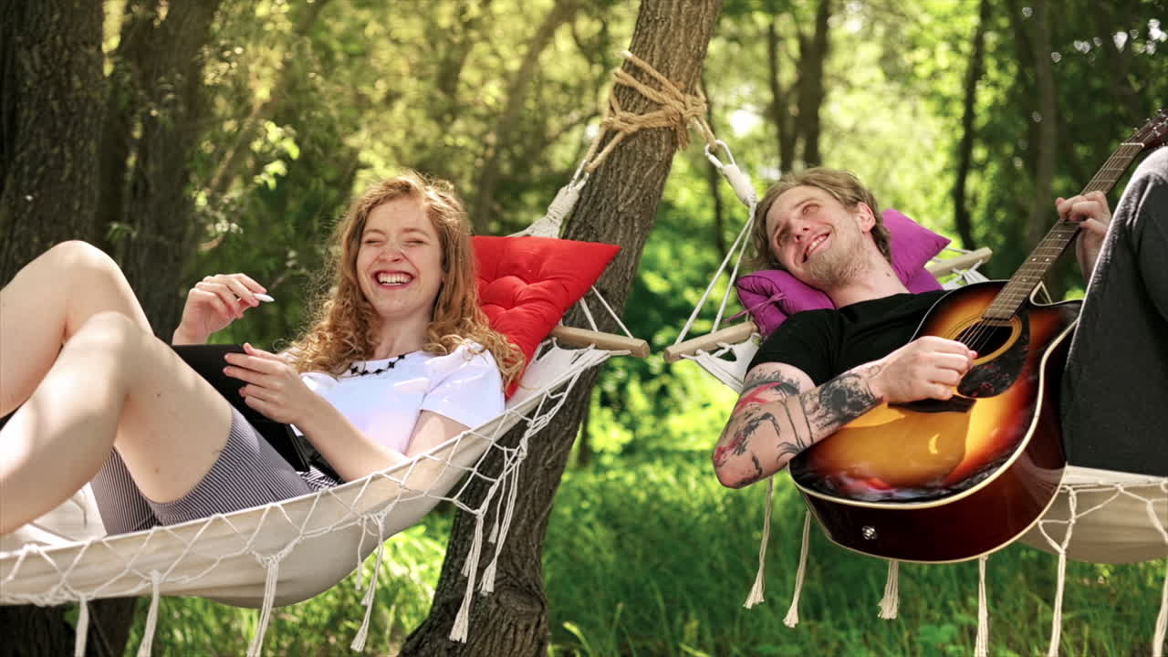 Couple lying on hammocks, talking and laughing. Young man with guitar, woman with tablet and stylus. Greenery around. Glamping. Slow motion