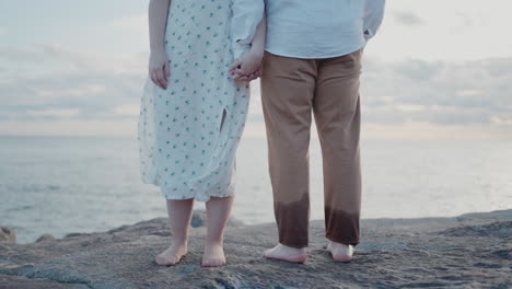 couple-standing-barefoot-on-rocks-near-the-ocean,-holding-hands-at-sunset